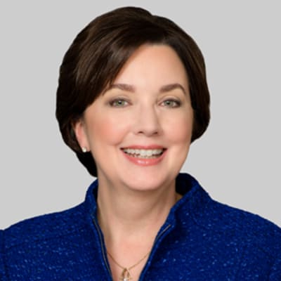 Carrie Penman, smiling, with short brown hair wearing a textured blue jacket against a plain gray background.