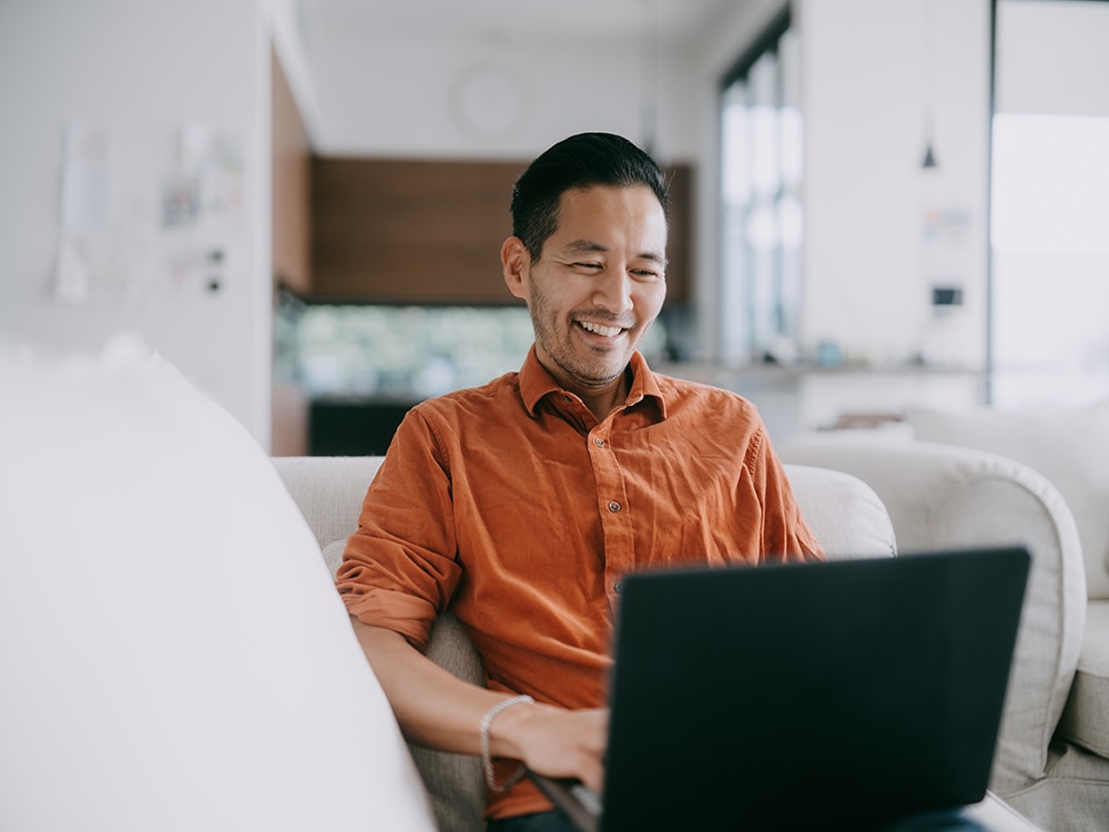 A man in an orange shirt smiles while using a laptop on a light-colored sofa in a bright, modern living room. The room features large windows and visible kitchen elements in the background.