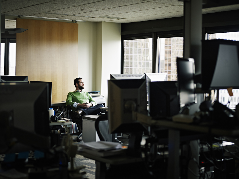 A man in a green sweater sits at a desk in a dimly lit office, looking out the window. The office is filled with computers and office supplies. Sunlight streams through the window, casting a soft glow over the scene.