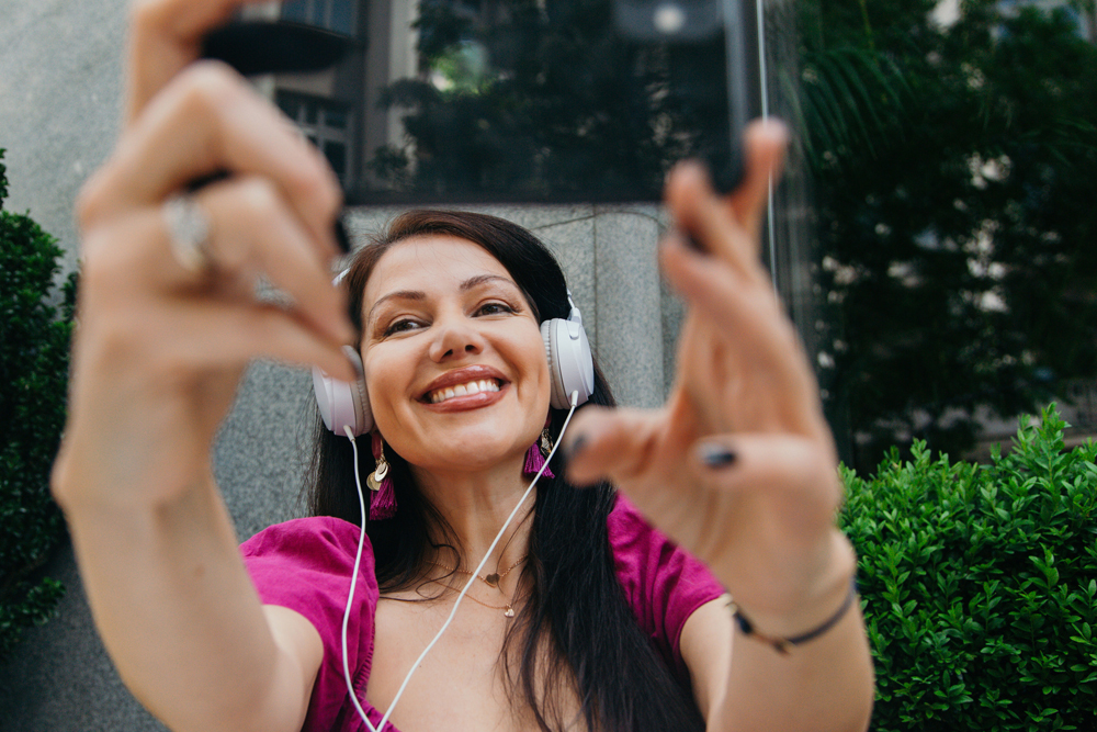 A woman with long dark hair wearing white headphones is taking a selfie. She is smiling and sitting outdoors, surrounded by greenery. She wears a bright pink top and appears joyful and relaxed.